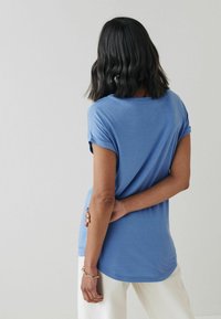 T-shirt blu a collo rotondo con maniche corte arrotolate, realizzato in tessuto morbido, vestibilità ampia e orlo leggermente curvo. Indossato con pantaloni bianchi.