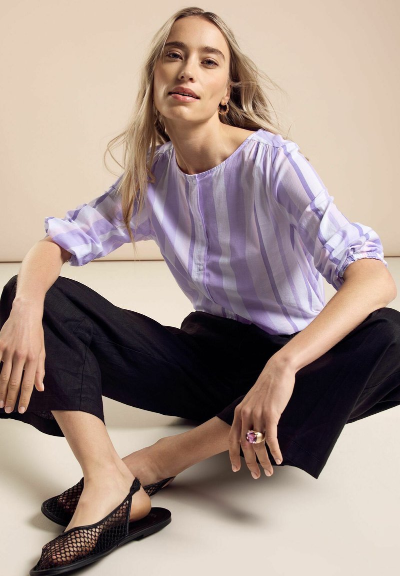 Blusa a righe viola chiaro con maniche a palloncino corte, abbinata a pantaloni neri a gamba larga e scarpe slingback nere in rete. Grande anello alla mano.