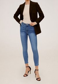 Blazer negro sobre una blusa beige de canalé, combinado con jeans ajustados de pitillo azul claro y tacones de tiras negros. Los jeans tienen un lavado desgastado y un dobladillo deshilachado.
