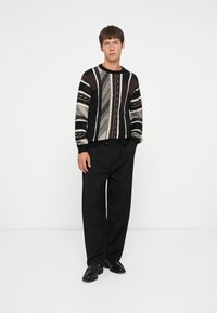 Sort og hvid mønstret sweater med teksturerede striber, båret med løse sorte bukser og sorte sko. Enkel baggrund.