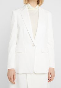 Blazer bianco sartoriale con revers a lancia, due tasche frontali e chiusura con un solo bottone. Tessuto liscio con silhouette strutturata.