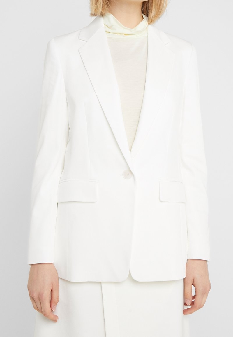 Blazer bianco sartoriale con revers a lancia, due tasche frontali e chiusura con un solo bottone. Tessuto liscio con silhouette strutturata.