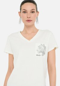 Camiseta de manga corta blanca con escote en V y un gráfico de leopardo en el bolsillo del pecho; hecha de una tela suave y ligera. Etiqueta de marca: LIU JO.
