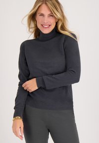 Donkergrijze coltrui in een gladde brei, met lange mouwen en een geribbelde zoom. Combinatie met donkere broek en een gouden armband.