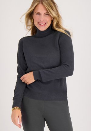 Donkergrijze coltrui in een gladde brei, met lange mouwen en een geribbelde zoom. Combinatie met donkere broek en een gouden armband.
