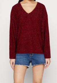 Haut à manches longues bordeaux en tissu texturé avec un décolleté en V, assorti à un short en denim effiloché, mettant en valeur un look décontracté.