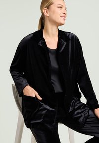 Black velvet jacket with a collar, buttoned front, and side pockets, paired with a black top. Smooth texture and loose fit accentuate comfort.