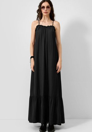 Femme portant une longue robe noire ample avec de fines bretelles et des lunettes de soleil, debout devant un fond uni gris clair.