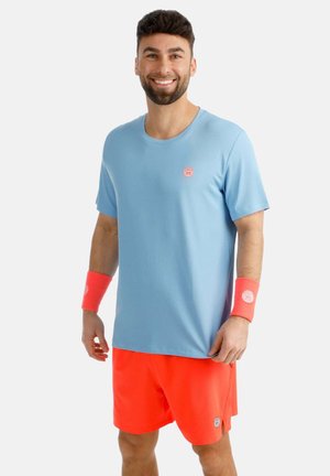 Uomo sorridente che indossa una t-shirt azzurro chiaro, pantaloncini arancioni brillanti e braccialetti arancioni abbinati, in piedi davanti a uno sfondo chiaro uniforme.