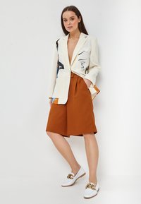 Blazer with abstract designs in white and colors, paired with brown shorts. Footwear is white with a decorative knot detail.