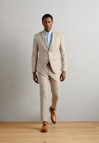 Completo beige chiaro con giacca su misura e pantaloni slim, abbinato a una camicia dress azzurro chiaro e scarpe in pelle marrone. Texture liscia.