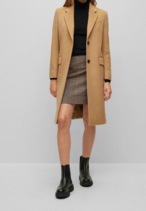 Vrouw draagt een camel-kleurige jas over een zwarte coltrui, een houndstooth-minirok en zwarte enkellaarzen tegen een effen lichte achtergrond.