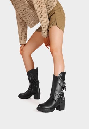 Ben iført sorte læderstøvler midt på læggen med spænder, kombineret med beige shorts og en beige strikket langærmet top, mod en hvid baggrund.