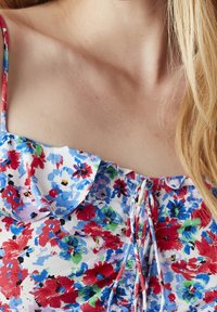 Haut à motifs floraux présentant une palette de couleurs bleue, blanche, rouge et verte, avec des bretelles froncées et un détail à nouer au niveau du col.