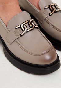 Grijze leren loafers met een stevige zwarte zool, voorzien van een goud-zwart kettingaccent over de wreef. Glad afgewerkt met stiksel details.