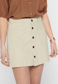 Gonna di cotone beige chiaro a taglio dritto, con chiusura anteriore a bottoni e due tasche applicate sul retro, dettagliate con bottoni neri.