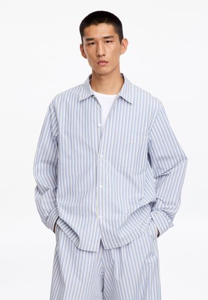 Jeune homme aux cheveux courts et noirs portant une chemise bleue et blanche à rayures verticales boutonnée et un pantalon ample assorti, les mains dans les poches.