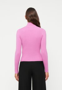 Frau mit langen braunen Haaren, die einen eng anliegenden, rosa gerippten Rollkragenpullover und schwarze Hosen trägt, von hinten vor einem schlichten Hintergrund gezeigt.