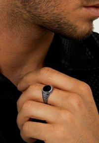 Schwarzer Steinsring mit silbernem filigranem Band, das feine Muster und eine polierte Oberfläche zeigt, getragen an einer Hand vor einem dunklen Hintergrund.
