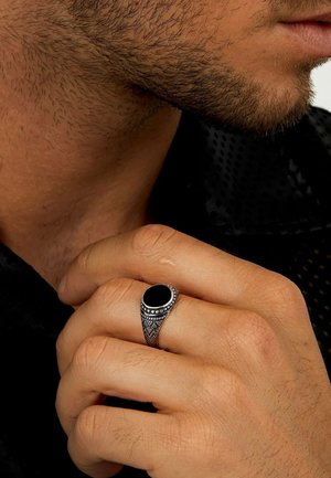 Silberner Ring mit einem ovalen schwarzen Stein, verziert mit filigranen Details und einem strukturierten Band. Hand liegt gegen ein dunkles Hemd.