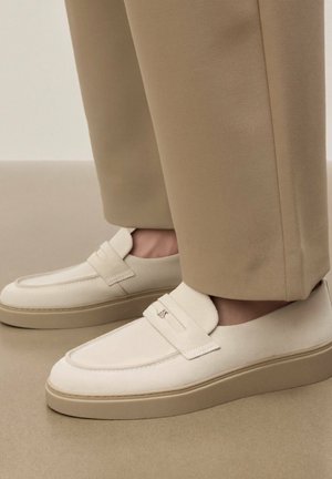 Cremefarbene Wildleder-Loafer mit dicken beigen Sohlen, getragen zu hellbraunen Hosen auf einem neutralen Boden und Hintergrund.