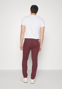 Jean slim bordeaux en mélange de coton, avec un design de poche arrière, porté avec un t-shirt blanc uni et des baskets de couleur claire.