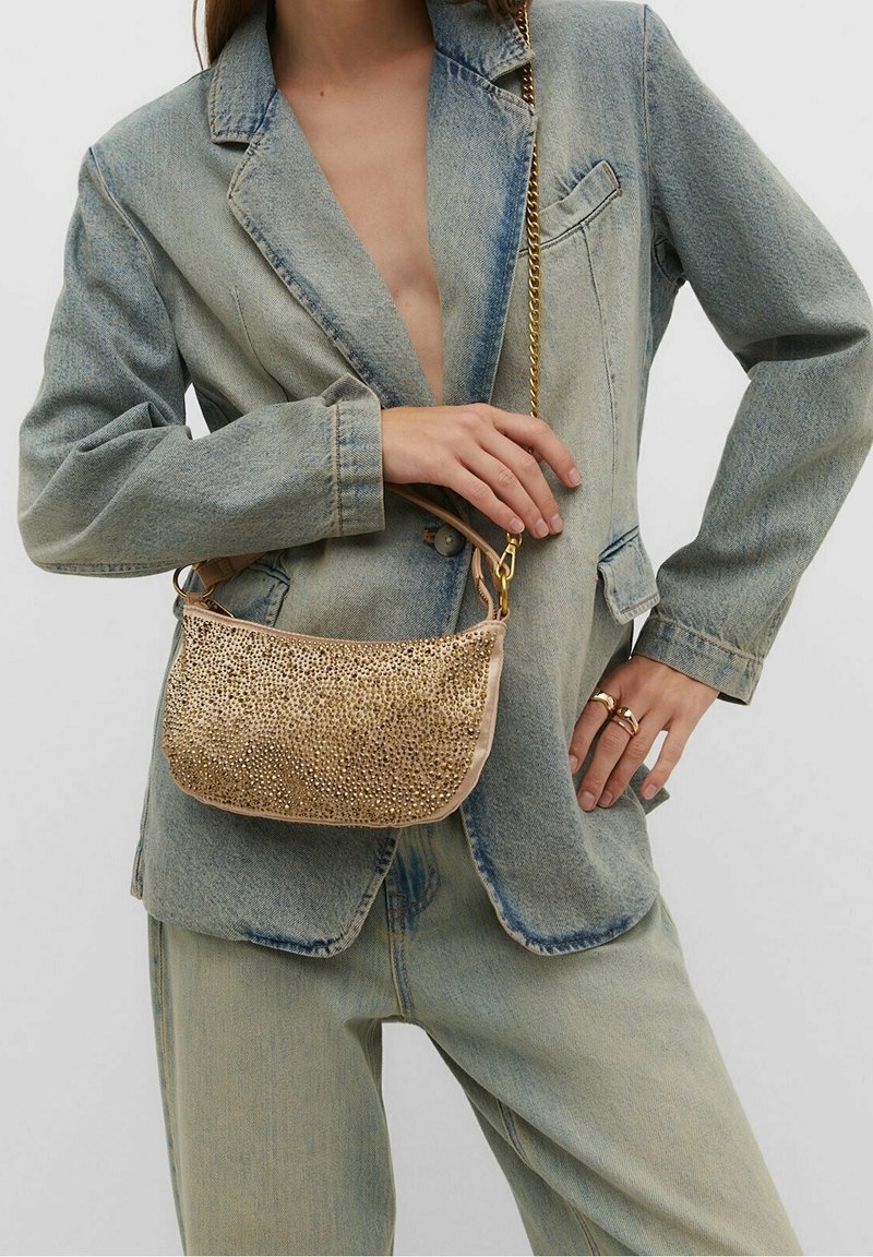 Femme portant un blazer et un pantalon en denim effet délavé, tenant un petit sac à main beige orné de strass dorés et une bandoulière chaîne dorée.