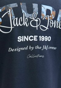 Navyblauwe katoenen T-shirt met grote bedrukte tekst: "STUD Jack & Jones," "SINCE 1990," en "Ontworpen door de J&J crew" in witte en metallic accenten.