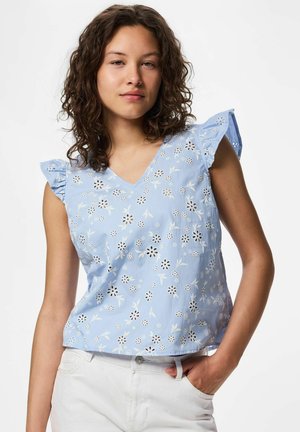 Mlajša ženska z kodrastimi lasmi, oblečena v svetlomodro cvetlično brezrokavnik in bele hlače, roka v žepu, na enobarvni podlagi.