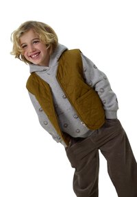 Braune gesteppte Weste über einem grauen Hoodie mit Smiley-Gesicht-Muster, kombiniert mit braunen Cord-Hosen. Das Outfit hat eine lockere Passform.