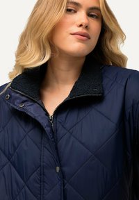 Giubbotto imbottito blu navy con zip frontale, dettagli con bottoni a pressione e un colletto nero testurizzato. Presenta un motivo a rombi e un tessuto elegante.