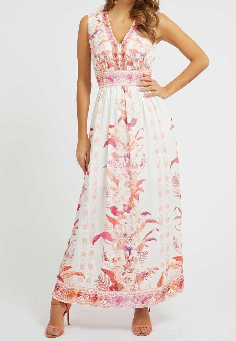 Femme portant une robe maxi blanche sans manches avec des motifs floraux roses et orange et des sandales à talons hauts, debout avec une main sur la hanche.