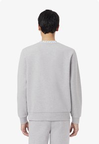 Sweatshirt gris avec un col rond, des poignets et un ourlet côtelés. Présente un col marqué avec "LACOSTE" en blanc. Tissu doux et texturé.