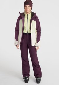 Die Skiausrüstung besteht aus einer lila Jacke mit cremefarbenen und grünen Akzenten, kombiniert mit passenden lila Hosen. Enthält einen schwarzen Gürtel und Schneestiefel.