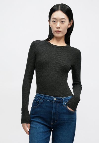 Γυναίκα με ίσια μαύρα μαλλιά, φορεί μια εφαρμοστή μακρυμάνικη πλεκτή μπλούζα σκουρόφαιου γκρι χρώματος και μπλε τζιν, με το ένα χέρι στην τσέπη, ουδέτερο φόντο.