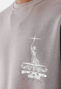 Sudadera gris claro con un gráfico central blanco de una estatua sosteniendo una antorcha, con el texto "PXP CAMBIA EL JUEGO" debajo.