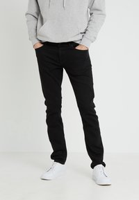 Jeans de mezclilla negra con corte ajustado, trabillas para cinturón y bolsillos frontales, combinados con una sudadera gris y zapatillas blancas. Textura suave, diseño casual.