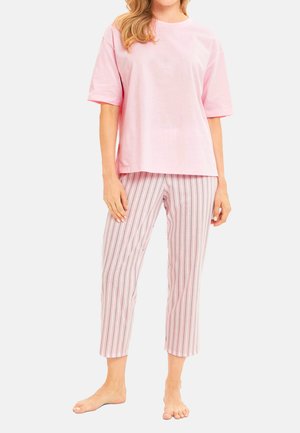 Frau trägt ein hellrosa kurzärmeliges Shirt und pink-weiß gestreifte kurze Hosen, steht barfuß auf einfarbigem Hintergrund.