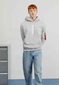 Felpa grigia chiara con tasca a marsupio frontale, etichetta con logo e tasca con zip rossa sulla manica, abbinata a jeans blu.