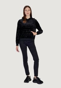 Sudadera negra con capucha y estampado de logo texturizado, mangas largas y ribete acanalado. Combinada con pantalones ajustados negros y zapatillas deportivas.