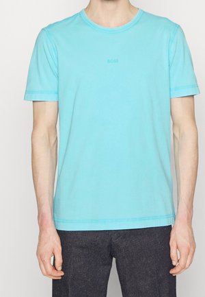 Mand iført en ensfarvet lyseblå T-shirt med korte ærmer og lille "BOSS"-logo på brystet, samt mørke bukser, stående foran en hvid baggrund.