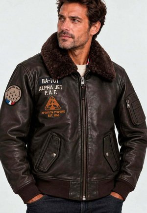 Homme portant une veste aviateur en cuir marron foncé avec col en peau lainée et écussons d'aviation, mains dans les poches, regardant légèrement vers la droite.