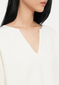 Λευκό πουλόβερ σε σχήμα V με απαλή υφή και χαλαρή εφαρμογή, που διαθέτει μια μικρή κοπή κοντά στο ντεκολτέ. Έμφαση στην απλότητα και την άνεση.