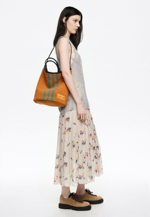 Vrouw in een bloemen- plisségemaakt rokje, mouwloze grijze top, tan wandelschoenen, en een oranje gestreepte tas met zwarte handvatten op haar schouder.