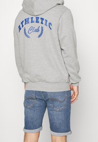 Šedá mikina s kapucí s modrým nápisem "ATHLETIC Club" a vavřínovým vzorem na zádech, doplněná džínovými šortkami s vyhrnutými manžetami a zdobenými zadními kapsami.