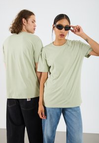 Light green oversized cotton t-shirts with short sleeves, worn by two models, paired with black and blue denim pants. Simple design, no patterns.