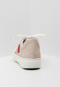 Beżowy tenisówka z płótna z białą gumową podeszwą, z czerwonym haftem na pięcie i białą satynową kokardą na górze. Prostota i minimalistyczny design.