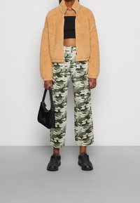 Veste zippée tan et moelleuse sur un crop top noir ; pantalon cargo vert camouflage avec poches ; baskets noires ; tenant un petit sac noir.