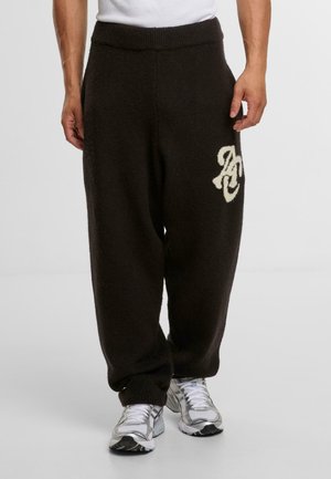 Brown fleece sweatpants featuring a ribbed waistband, side pockets, and a cream-colored logo on the left leg. Relaxed fit.