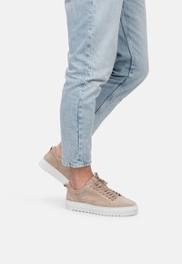 Zapatillas de ante beige con una superficie texturizada, suela de goma blanca y cordones planos. Llevadas con jeans ajustados de color azul claro, enfatizando el calzado.